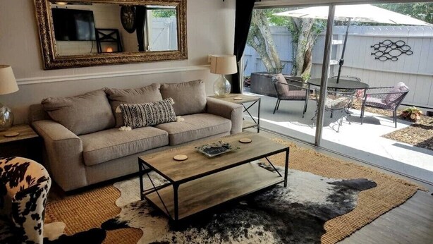 Un salon comprenant un canapé, une table basse et un grand miroir. Une porte-fenêtre coulissante donne accès à un patio extérieur aménagé avec une table et des chaises.