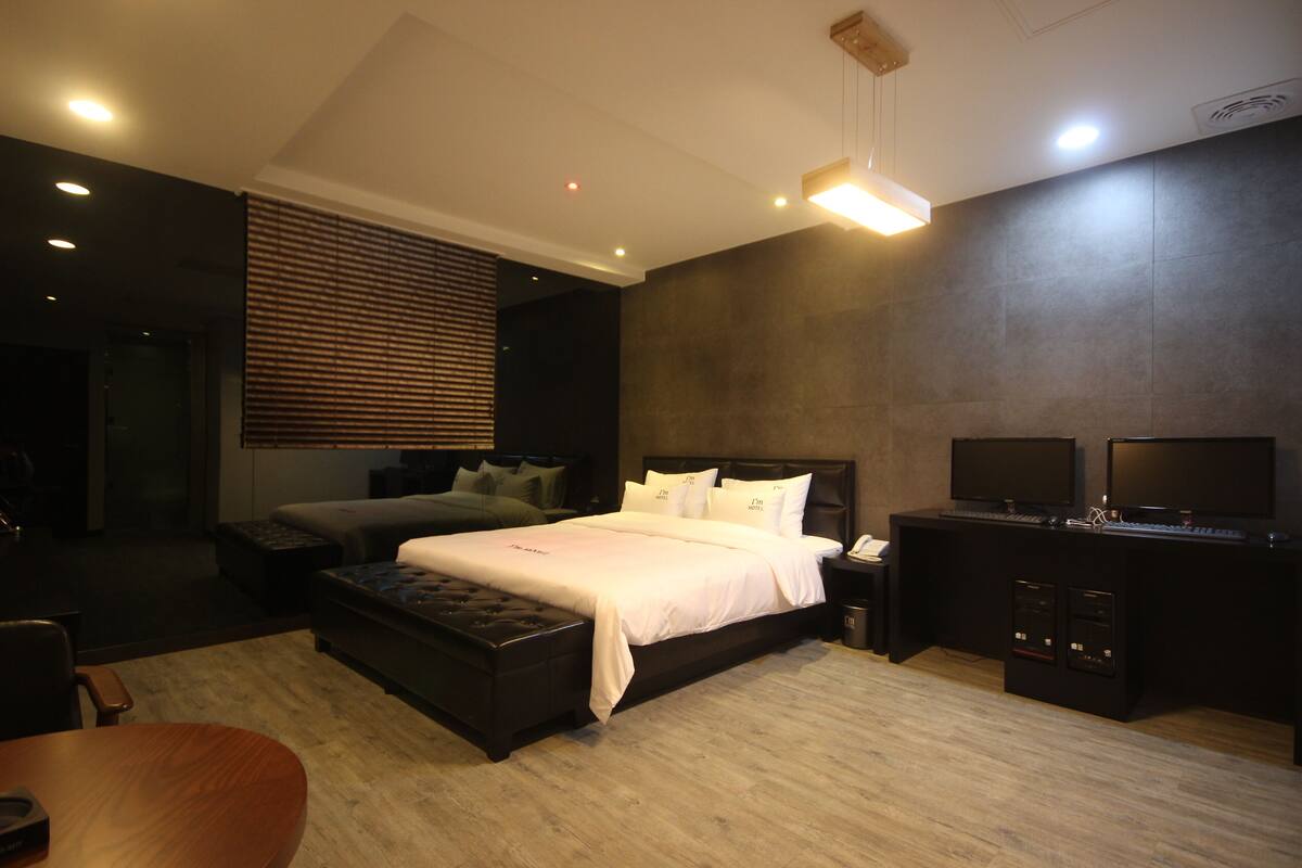 A modern hotel room with a large bed, a desk with two monitors, and a seating area with a sofa and ottoman.