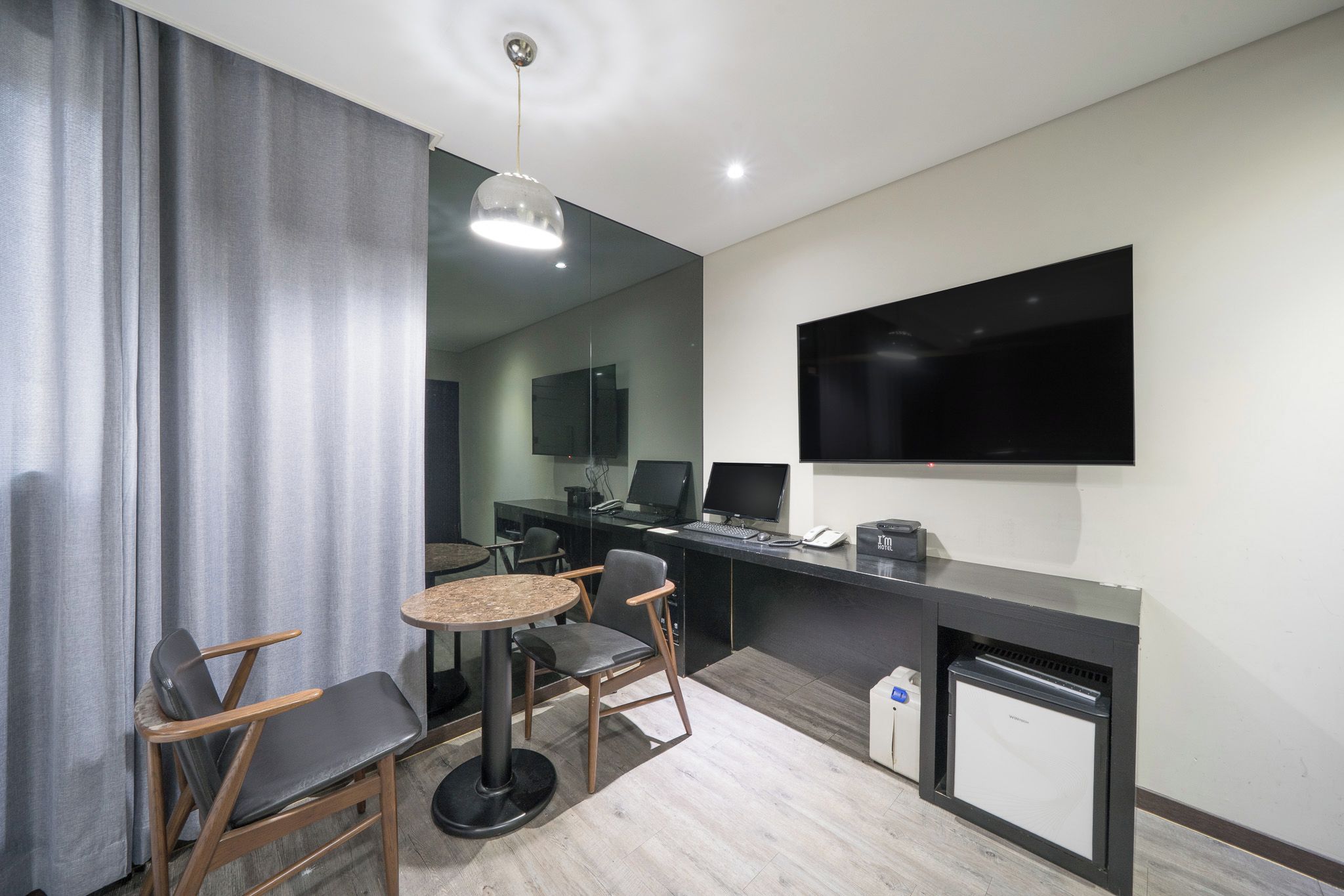 A modern hotel room with a flat-screen TV mounted on the wall, a desk with a computer, and a seating area with a round table and chairs.