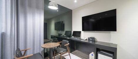 A modern hotel room with a flat-screen TV mounted on the wall, a desk with a computer, and a seating area with a round table and chairs.