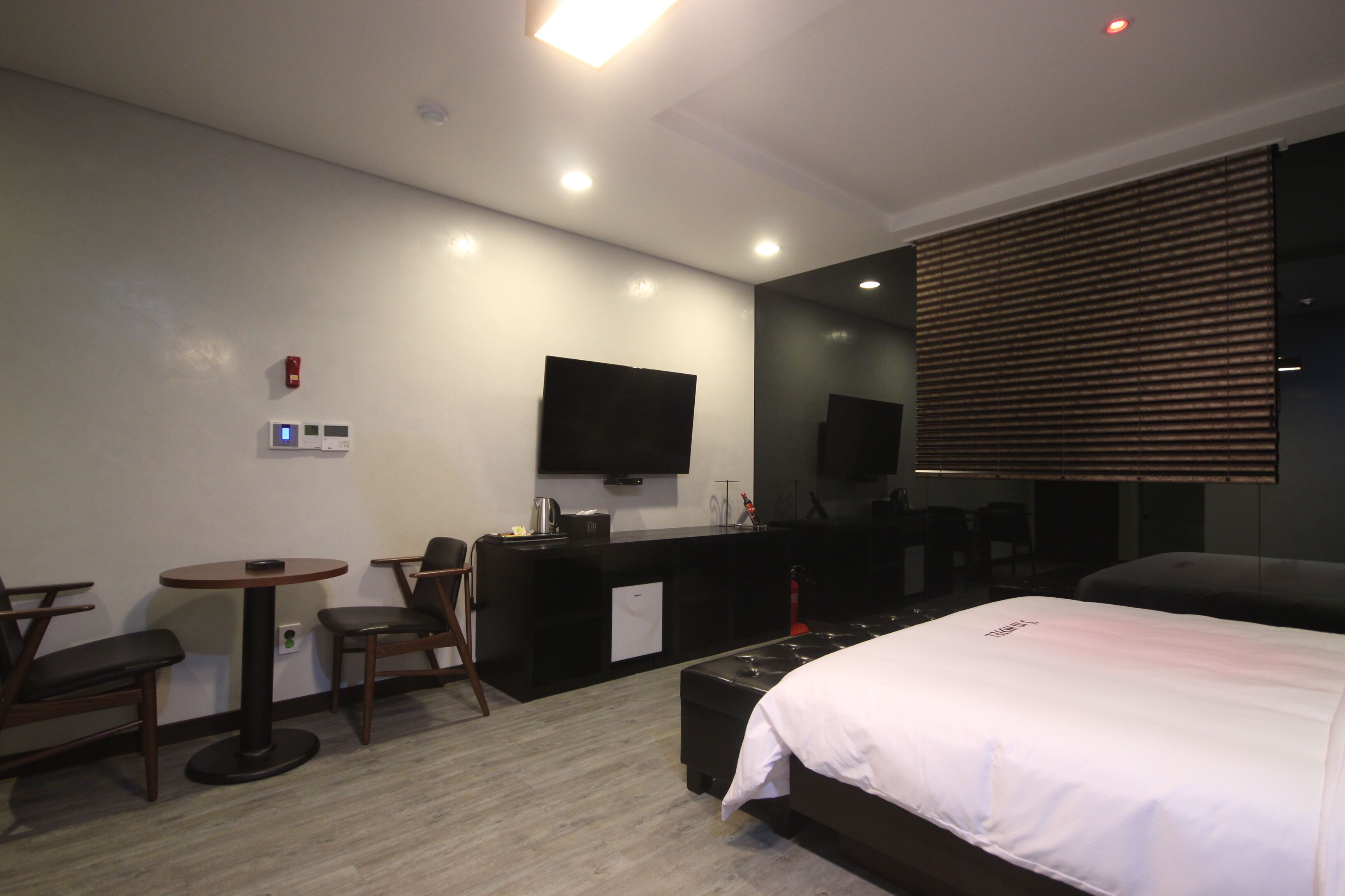 A hotel room with a bed, a desk, a chair, a small table, a TV, and a window with blinds.