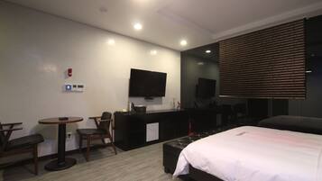 A hotel room with a bed, a desk, a chair, a small table, a TV, and a window with blinds.