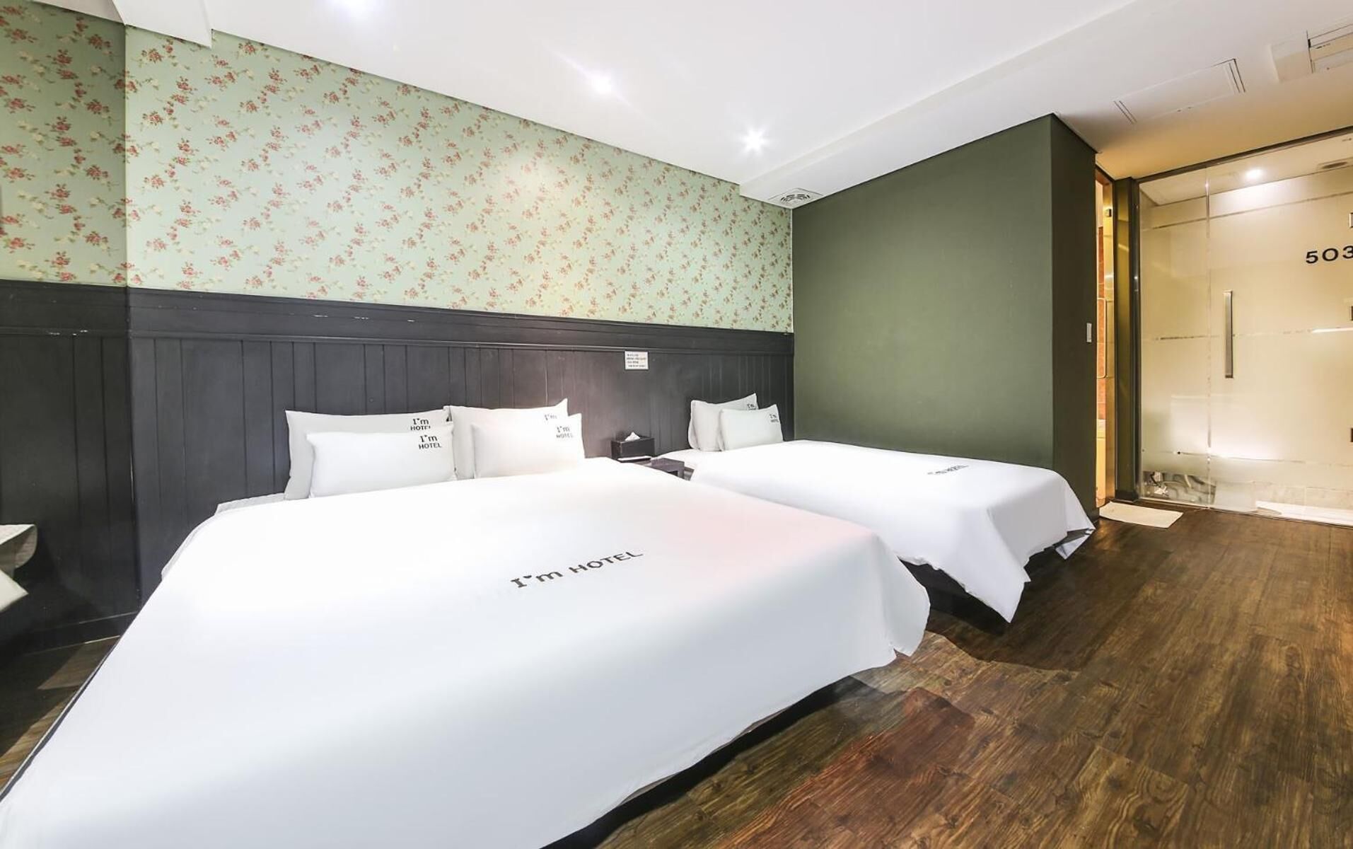 Habitación de hotel con dos camas, papel tapiz floral, cabecero de madera oscura y puerta de ducha de vidrio.