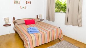 A bedroom with a bed, a window with curtains, a wall with framed pictures, and a small table with a lamp. - Pousada Jardim da Nova Era (Alto Paraíso de Goiás)