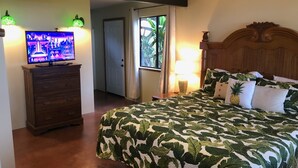 A hotel room with a bed, a television on a dresser, a window with curtains, and a door leading to another room. - 2 Bedroom Kona Coffee Farm with Ocean Views in Holualoa (Holualoa)