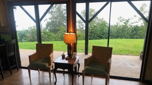 A room with two wicker chairs, a small table with a lamp, and a view of a green garden through large windows. - 2 Bedroom Kona Coffee Farm with Ocean Views in Holualoa (Holualoa)
