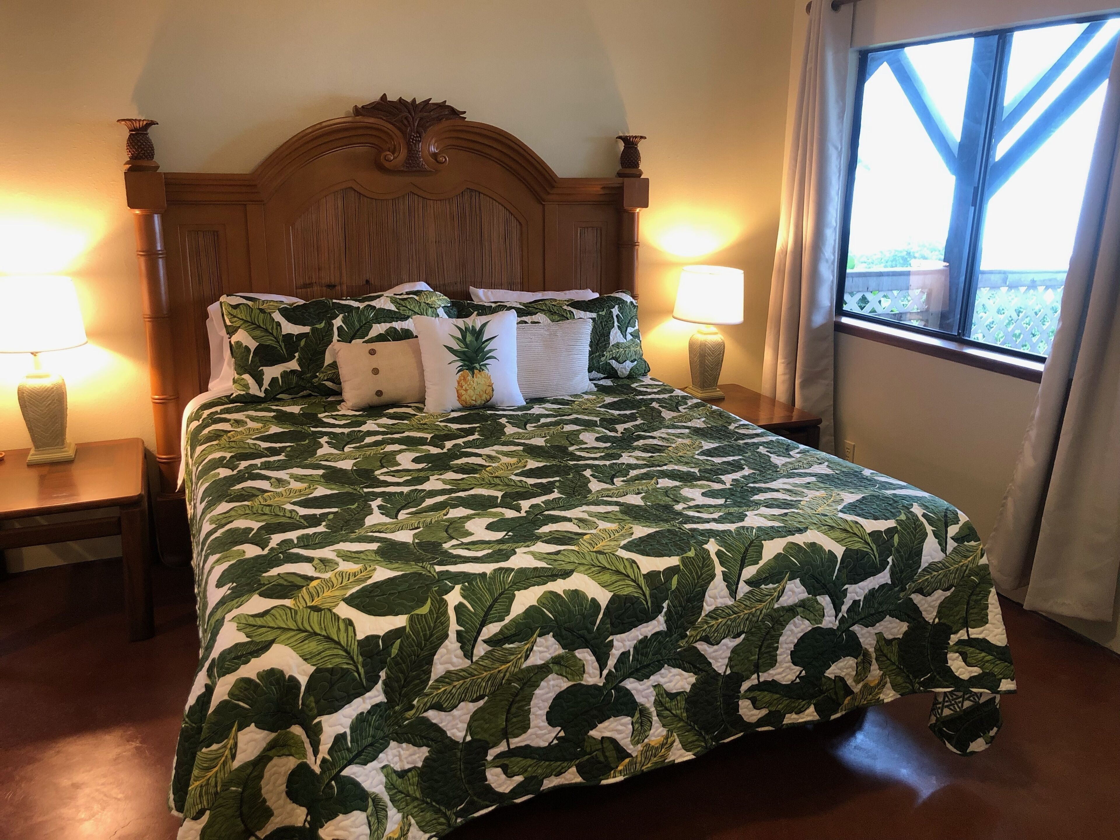 Cama com colcha estampada com folhagem tropical, com duas mesas de cabeceira com luminárias, cabeceira de madeira e uma janela com cortinas.