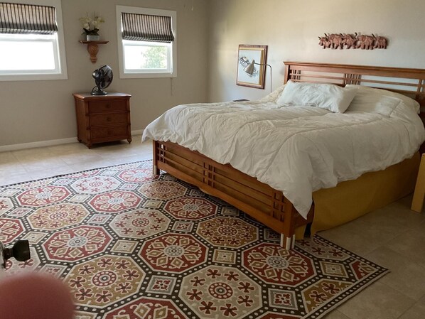 Une chambre avec un lit en bois, un tapis à motifs, deux fenêtres équipées de stores, une table de chevet et une sculpture d’éléphant fixée au mur.