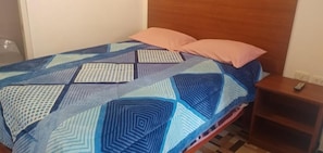 A bed with a geometric patterned bedspread, a wooden headboard, and a nightstand with a remote control. - Hospedaje San Pedro II (Cusco)
