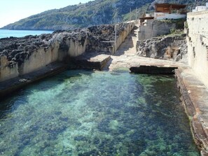 Une piscine en bord de mer, à l’eau claire, entourée de murs de pierre et de marches descendant vers l’eau.