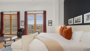 A modern bedroom with a large bed, a sofa, a glass table, and a view of the city through large windows. - The Axis - Condo on Dickson St - Walk to UofA (Fayetteville)