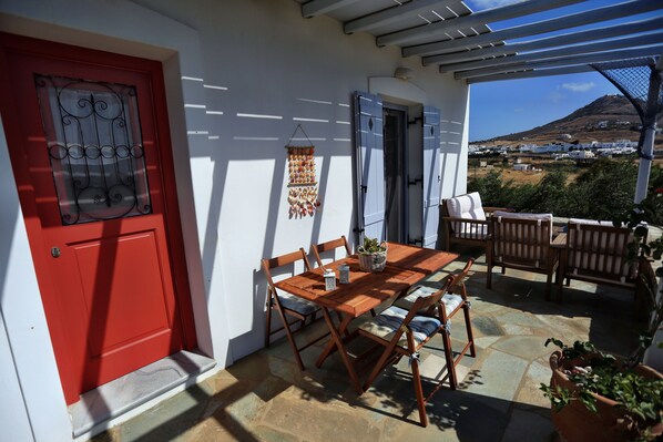 Une terrasse couverte avec une table et des chaises en bois, une porte rouge et une vue sur une colline.