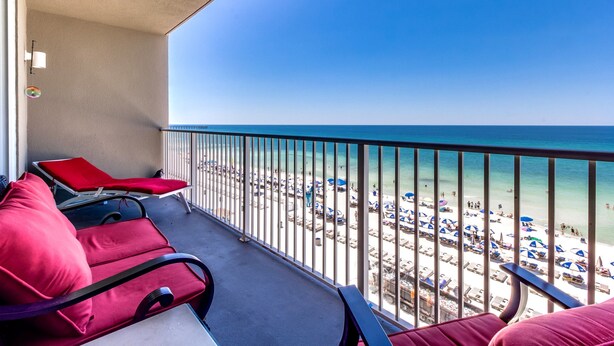 A balcony with red chairs and a table, overlooking a beach with umbrellas and people. - Tidewater by Forehand Rentals (Panama City Beach)