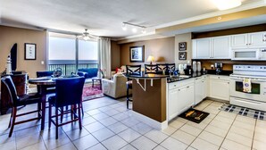 A modern kitchen with white cabinets, a central island, and a dining area with blue chairs. - Tidewater by Forehand Rentals (Panama City Beach)