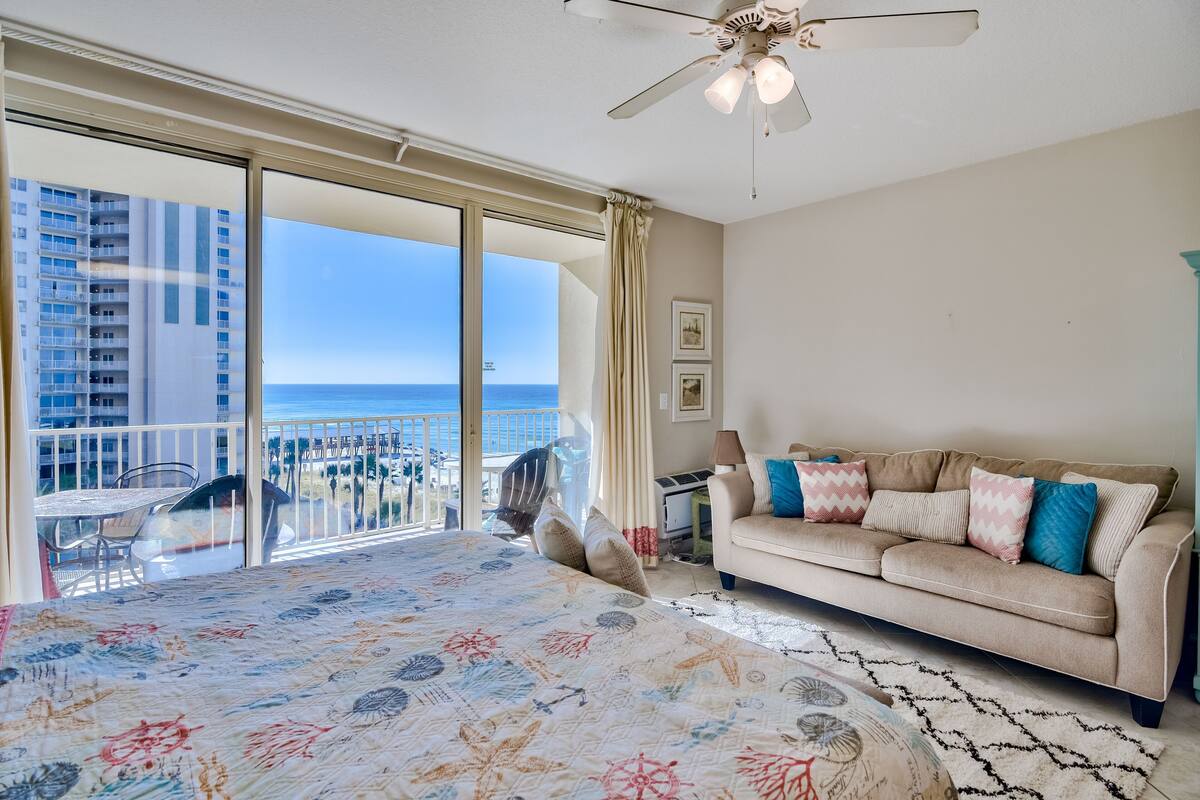 A bedroom with a large bed, a beige sofa, and a balcony with a view of the ocean.