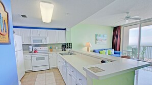 A modern kitchen with white cabinets, a microwave, and a stove. There is a dining area with a blue couch and a view of the ocean through large windows.