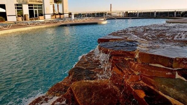 A swimming pool with a rocky edge, a building with balconies, and a clear sky.