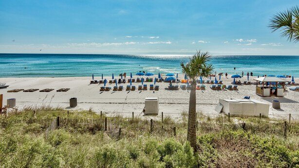 A beach with rows of sun loungers and umbrellas, a lifeguard tower, and a clear view of the ocean. - Celadon: Unit 102 by Forehand Rentals (Panama City Beach)