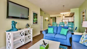 A living room with a blue sofa, white entertainment unit, and a green side table. - Celadon: Unit 102 by Forehand Rentals (Panama City Beach)