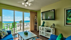 A living room with a view of the beach, a ceiling fan, a flat-screen TV, and a white console table. - Celadon: Unit 102 by Forehand Rentals (Panama City Beach)