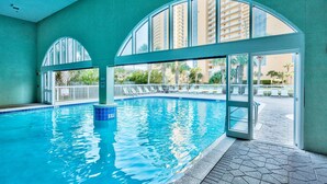 An indoor swimming pool with clear glass walls, a tiled floor, and a view of high-rise buildings in the background. - Celadon: Unit 102 by Forehand Rentals (Panama City Beach)