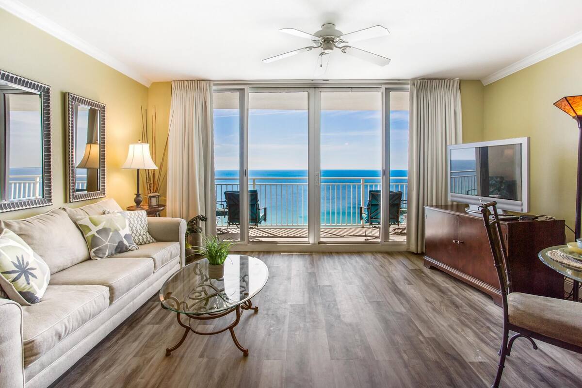 A living room with a sofa, coffee table, TV, and a balcony with a view of the ocean.