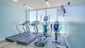 Et fitnessrum med løbebånd, elliptiske træningsmaskiner og en motionscykel, med et stort vindue der giver udsigt til havet.