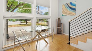 Un interior moderno con una mesa de comedor de cristal, sillas transparentes, una escalera con barandillas negras y vistas a la calle a través de amplios ventanales.