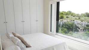 Een slaapkamer met een bed, nachtkastje, lamp en raam met uitzicht op groen en rotsen.