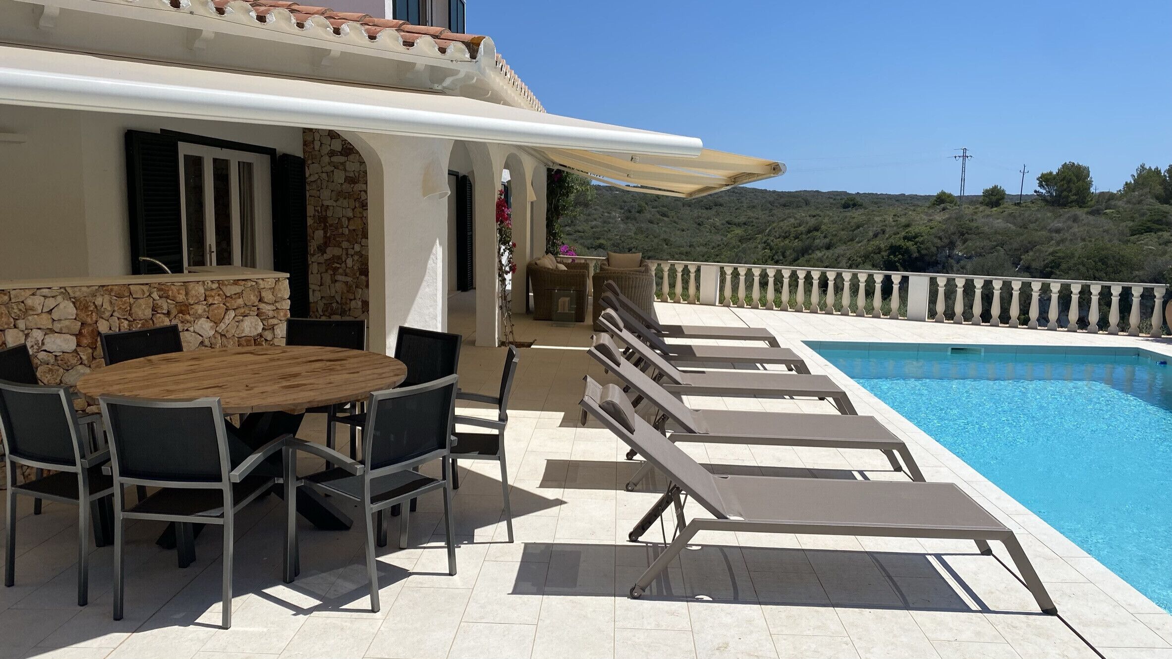 An outdoor pool area with lounge chairs, a dining table with chairs, and a stone wall.