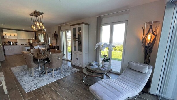 Ein modernes Wohnzimmer mit einer Couch, einem Esstisch und Stühlen, einer Küche im Hintergrund und großen Fenstern mit Blick.