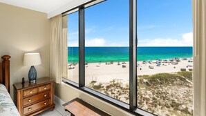 Quarto com uma grande janela com vista para a praia, cômoda de madeira e uma luminária.