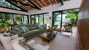 A spacious living area with a green sofa, white armchairs, and a wooden coffee table. Large windows offer views of a lush garden and a pool.