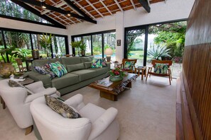 A spacious living area with a green sofa, white armchairs, and a wooden coffee table. Large windows offer views of a lush garden and a pool. - Pousada Chácara Aracê (Petrópolis)