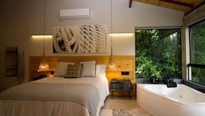 A bedroom with a large bed, a wooden headboard, a wall-mounted air conditioner, a bedside lamp, a window with a view of greenery, and a bathtub. - Pousada Chácara Aracê (Petrópolis)
