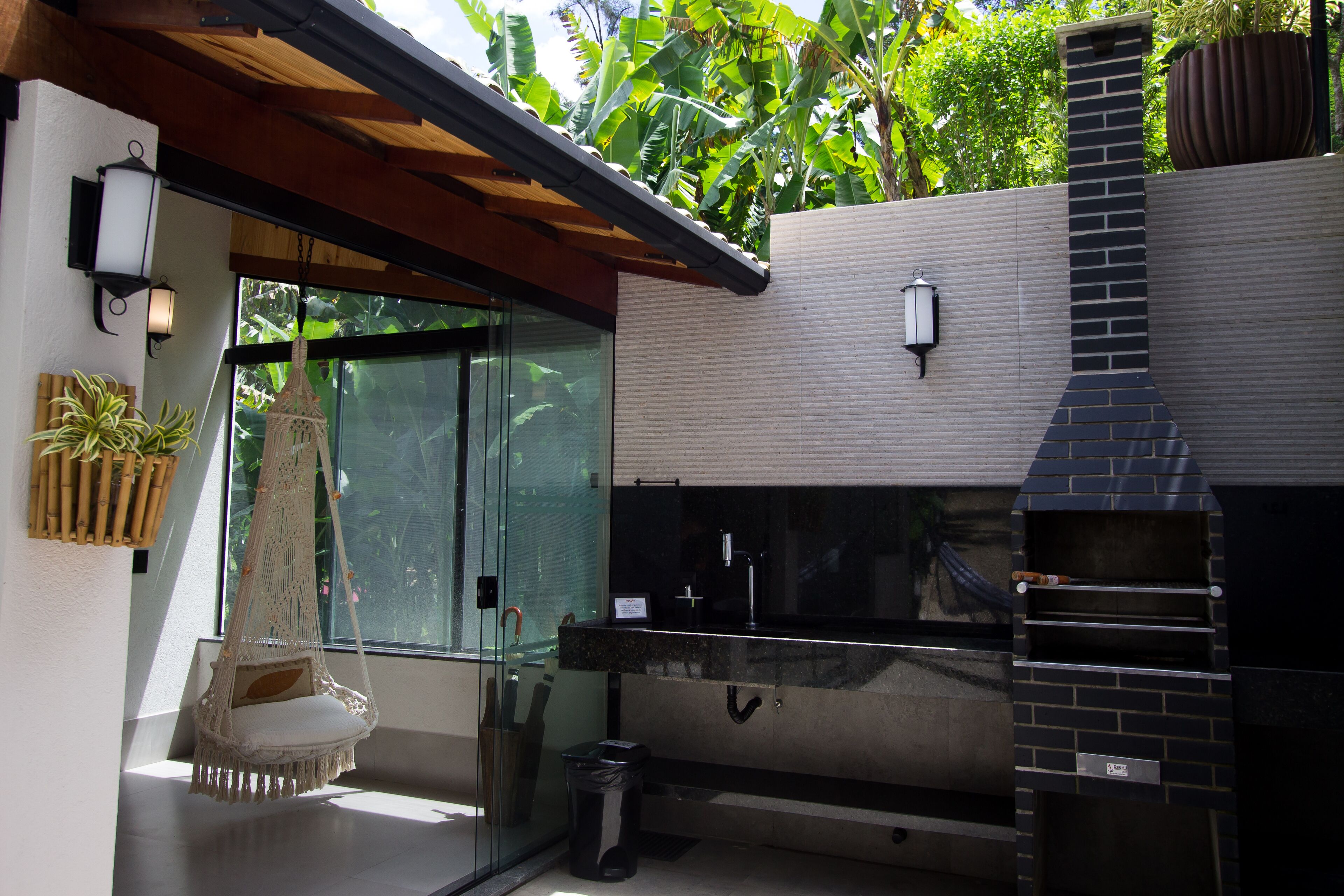 A modern outdoor kitchen with a brick oven, a hanging chair, and a glass door leading to a lush garden.