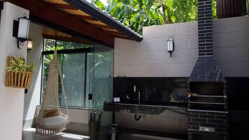 A modern outdoor kitchen with a brick oven, a hanging chair, and a glass door leading to a lush garden.
