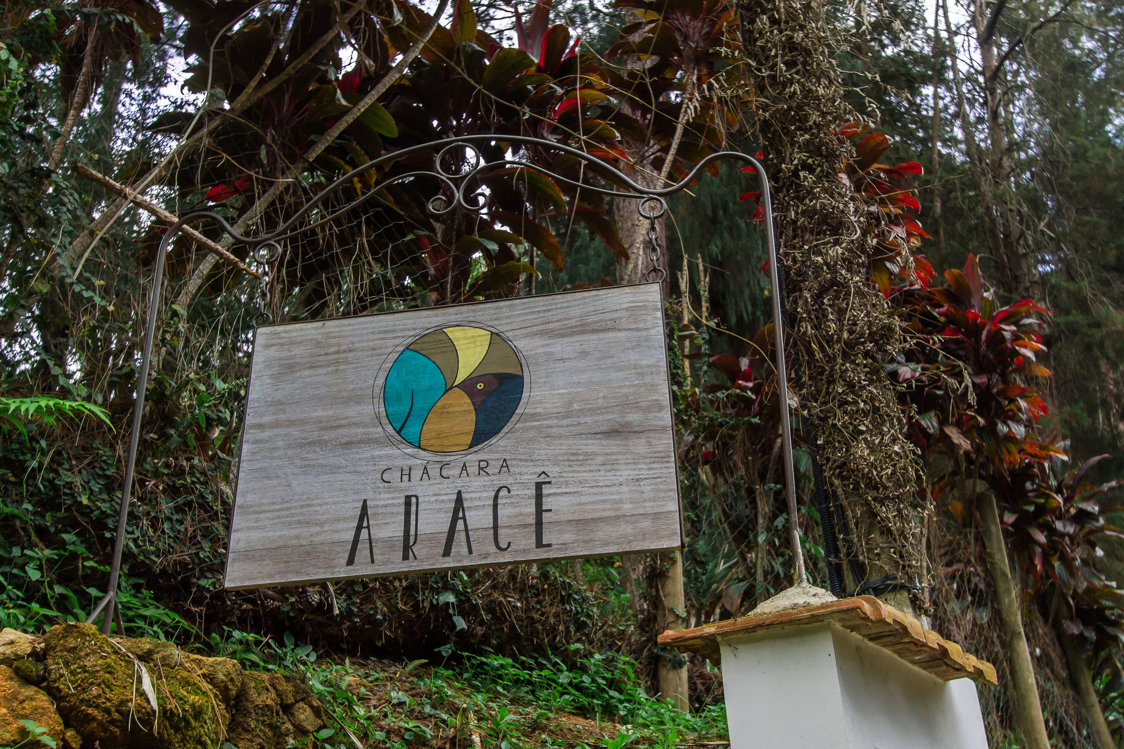 A wooden sign with a colorful logo and the text "Chacara APACE" against a backdrop of lush greenery and a metal structure.