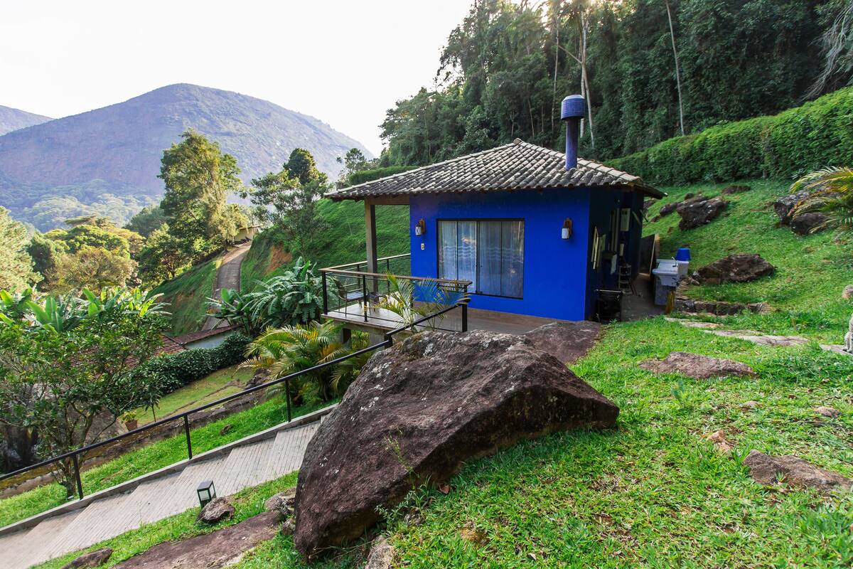 Um pequeno edifício azul com varanda, rodeado por vegetação exuberante e montanhas ao fundo.