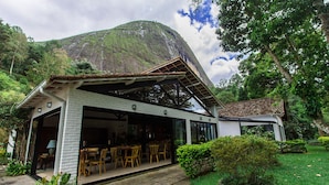 A modern building with a covered patio area, surrounded by lush greenery and a prominent mountain in the background. - Pousada Chácara Aracê (Petrópolis)