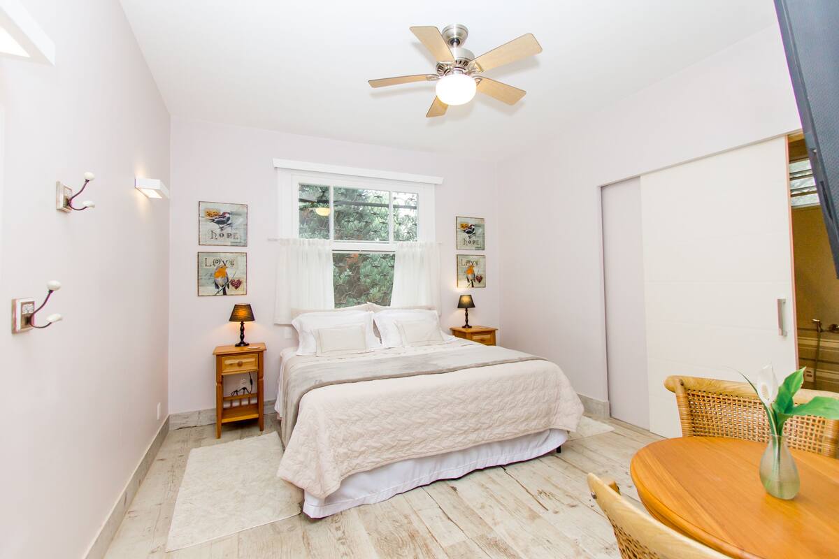 Um quarto com cama, mesas de cabeceira, ventoinha no teto, janela e uma mesa de jantar com cadeiras.