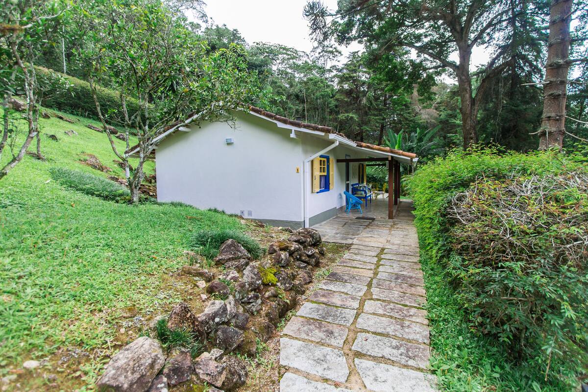 Um pequeno edifício branco com porta azul, rodeado por vegetação exuberante e um caminho de pedra.