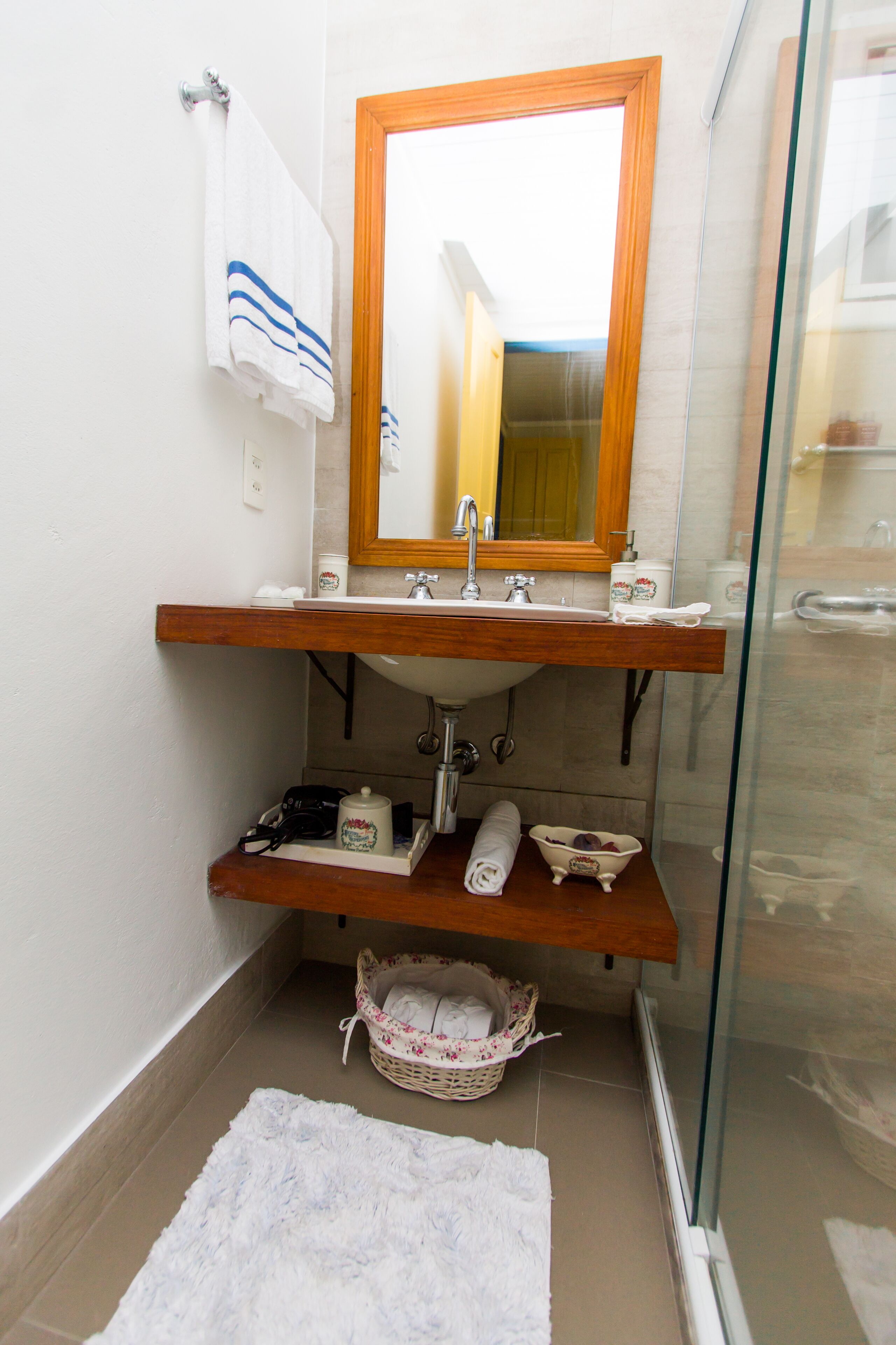 Un baño con tocador de madera, lavamanos, espejo, toallero, cabina de ducha de vidrio y una cesta de mimbre.