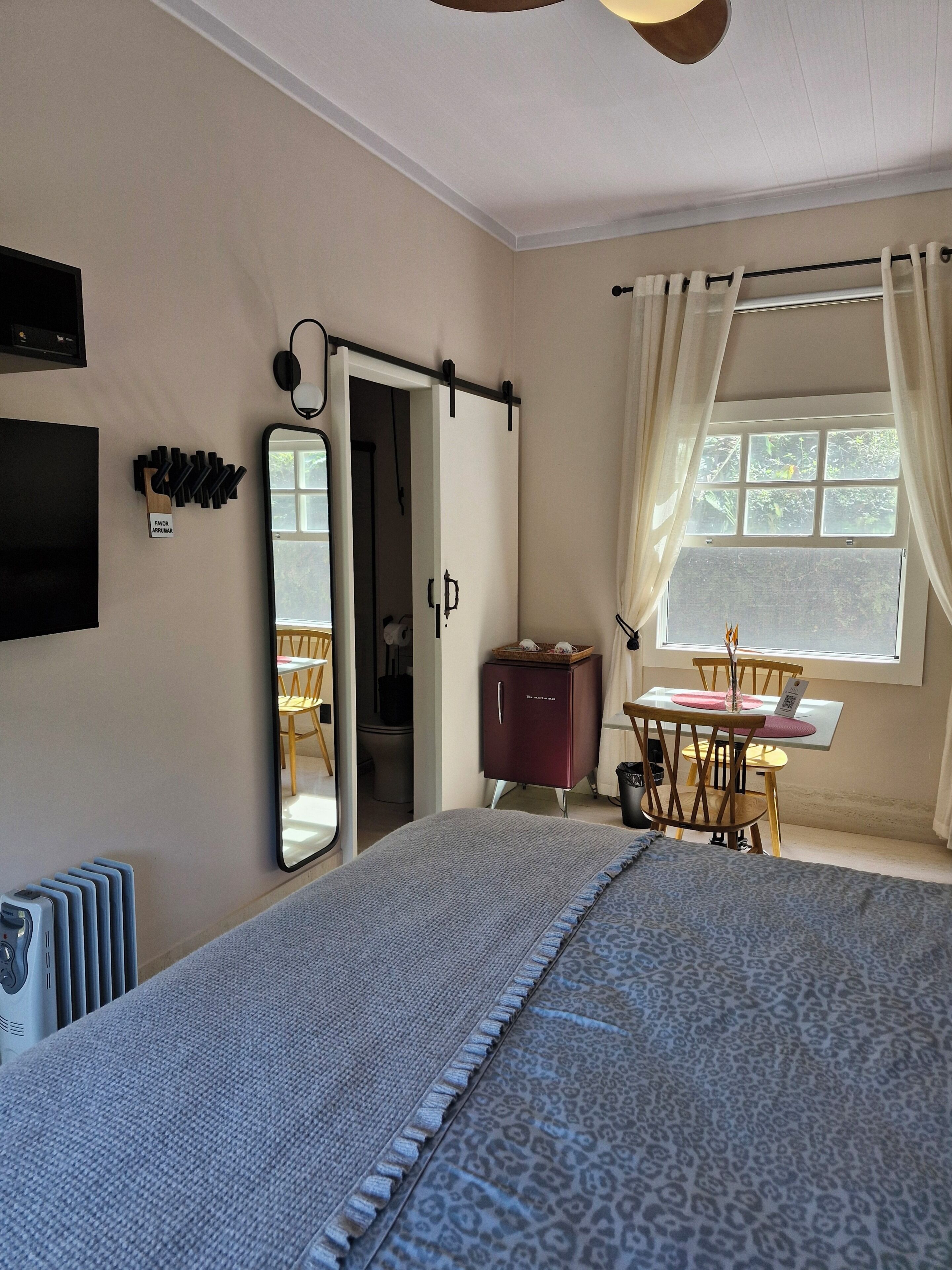 A bedroom with a bed, a desk, a chair, a radiator, a mirror, and a window with curtains.