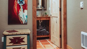 A hallway with a wooden door leading to a bathroom, a painting of a cow on the wall, and a small wooden cabinet with drawers. - 1- Bedroom 1- Bathroom Ranch Guest house. (Ellensburg)