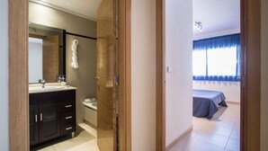 A hotel room with a bathroom and a bedroom. The bathroom has a sink, mirror, and towel rack. The bedroom has a bed, window, and curtains.