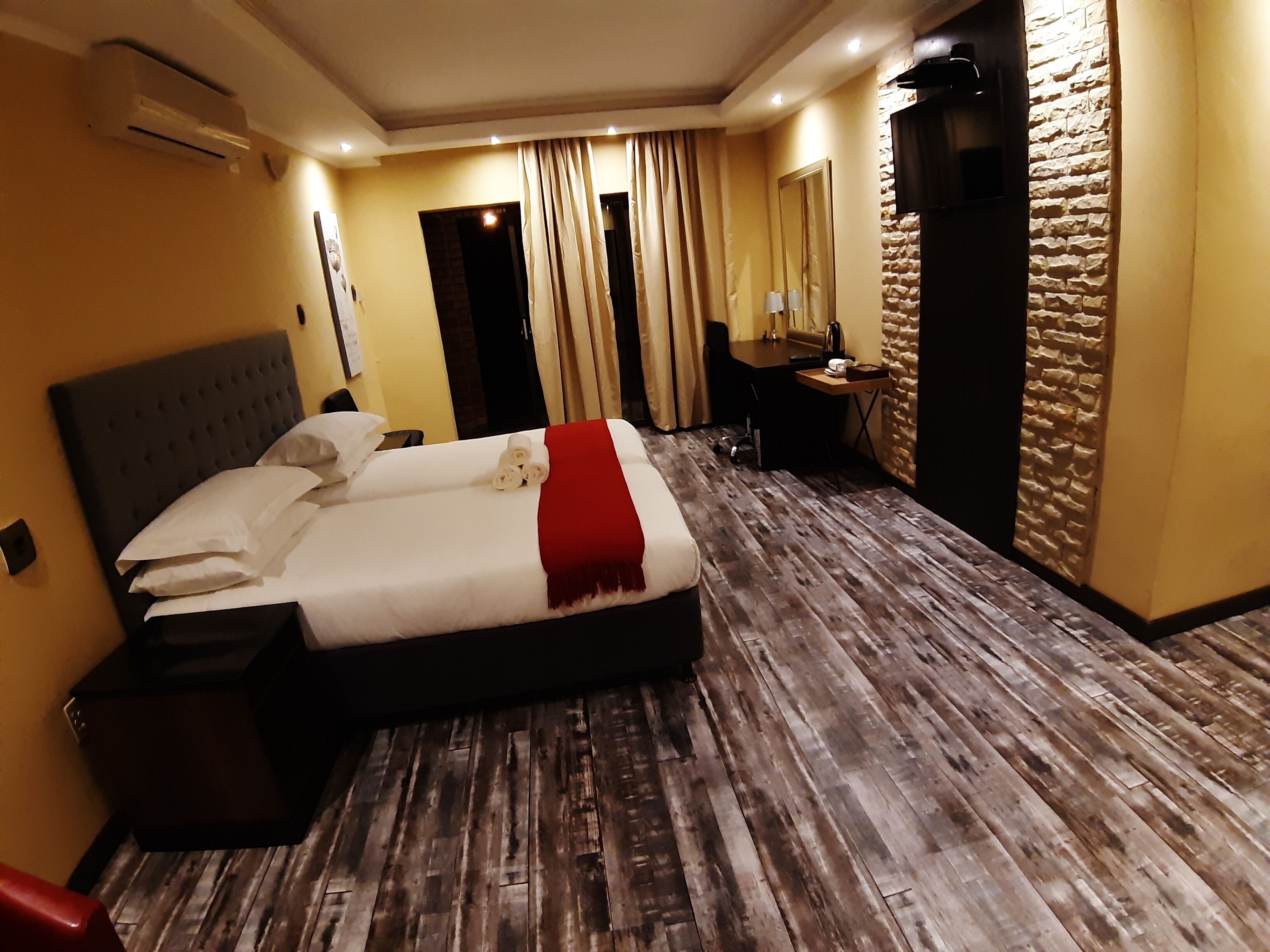 A hotel room with a large bed, a desk, a chair, an air conditioning unit, and a stone wall.