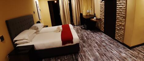 A hotel room with a large bed, a desk, a chair, an air conditioning unit, and a stone wall.