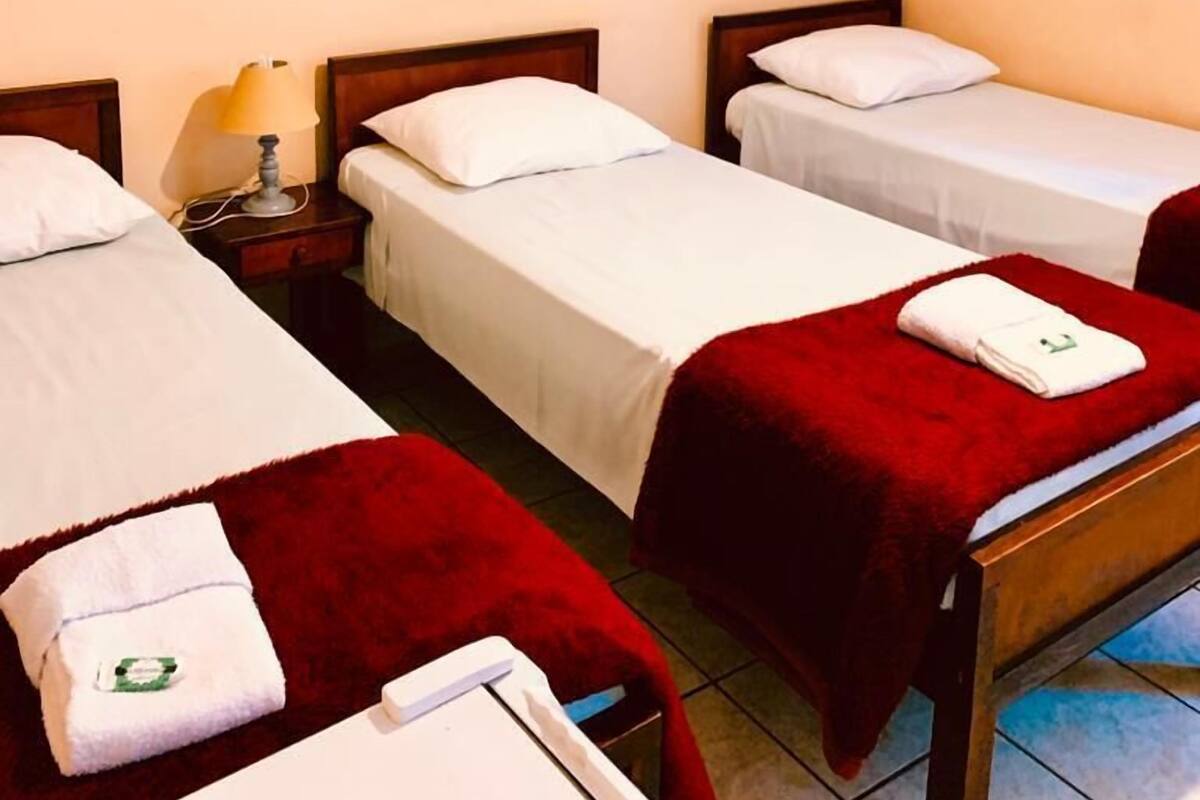A hotel room with two single beds, each with a red blanket and white pillows. There is a nightstand with a lamp between the beds.