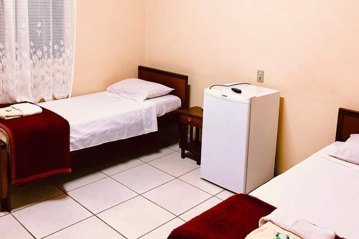 A hotel room with two single beds, a small desk, a refrigerator, and a window with a curtain.